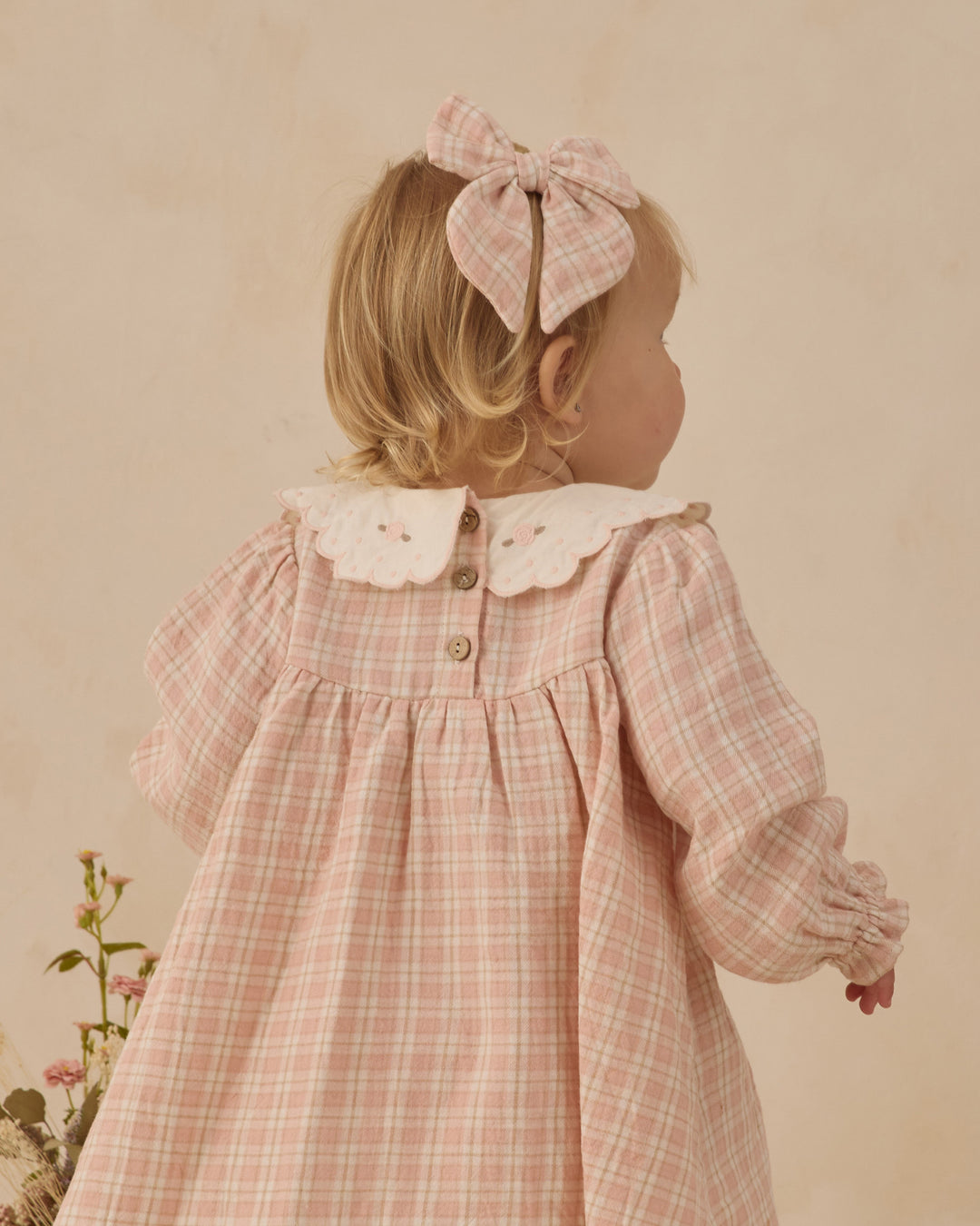 Young girl wearing a pink checkered dress with a large bow in her hair against a beige background