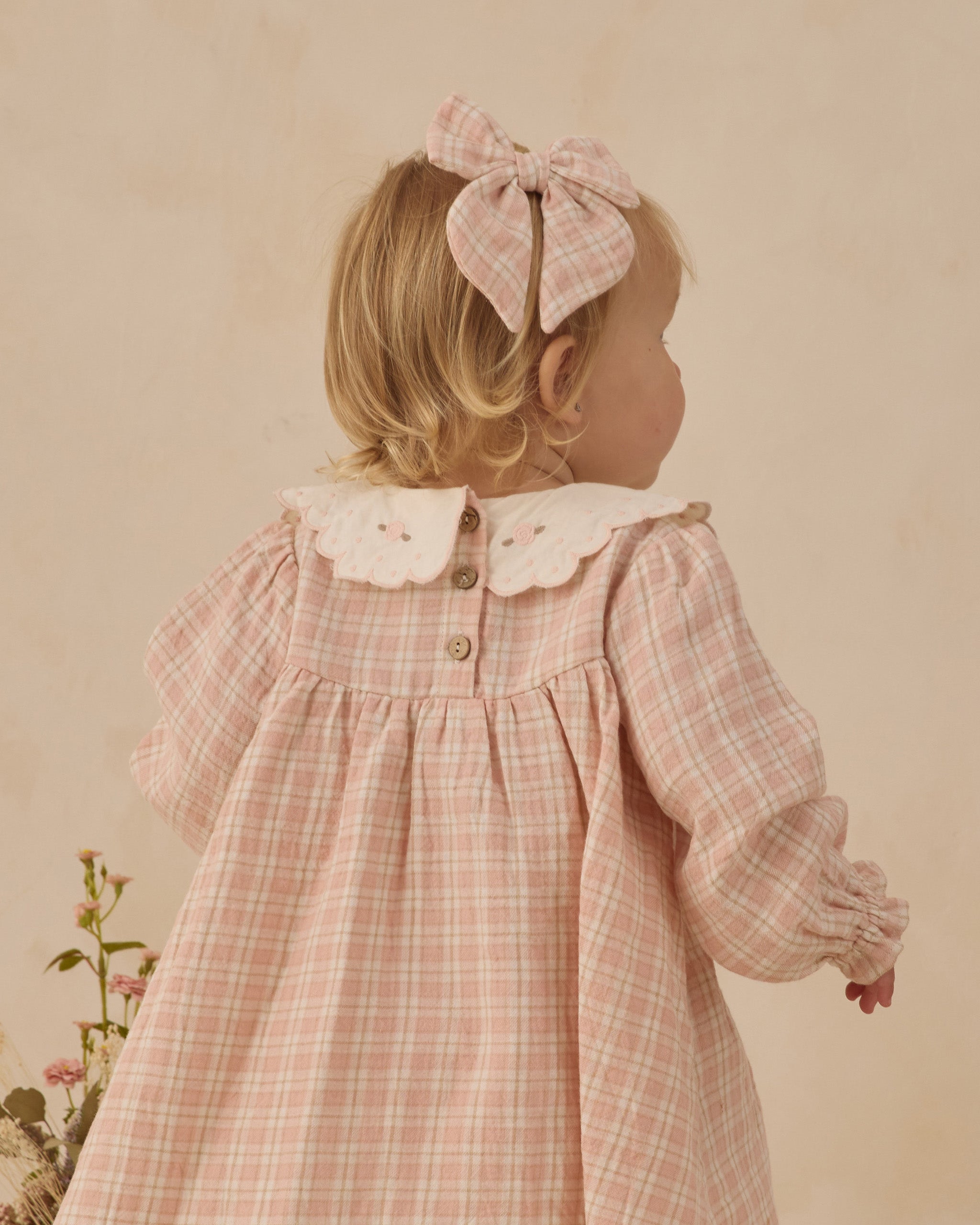 Young girl wearing a pink checkered dress with a large bow in her hair against a beige background