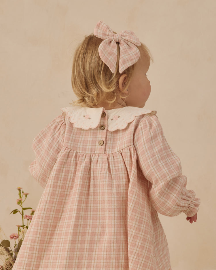 Young girl wearing a pink checkered dress with a large bow in her hair against a beige background